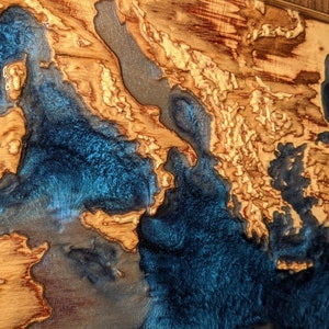 K&ouml;nnte beinhalten: Ein Wandkunstst&uuml;ck aus Holz mit einer blauen Harzeinlage. Das Harz ist so geformt, dass es an eine Karte oder ein Gew&auml;sser erinnert. Das Holz ist hellbraun mit einer nat&uuml;rlichen Maserung.