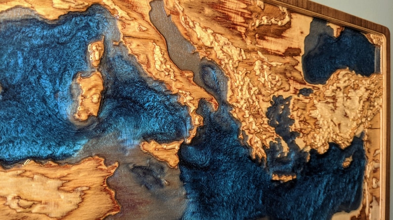 K&ouml;nnte beinhalten: Ein Wandkunstst&uuml;ck aus Holz mit einer blauen Harzeinlage. Das Harz ist so geformt, dass es an eine Karte oder ein Gew&auml;sser erinnert. Das Holz ist hellbraun mit einer nat&uuml;rlichen Maserung.