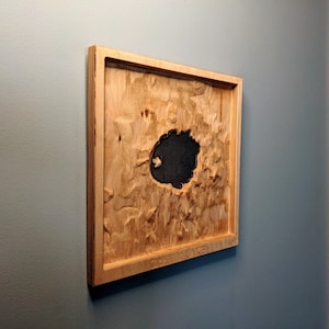May include: A wooden wall art piece with a carved out lake shape. The lake is painted black and the surrounding wood is a light brown colour. The text "CRATER LAKE" is engraved at the bottom of the piece.