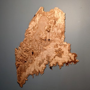 May include: A wooden map of the state of Maine, with a natural wood grain pattern and a light brown color.