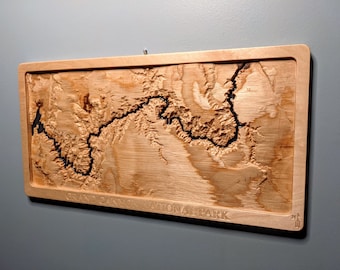 May include: A wooden wall art piece featuring a carved map of the Grand Canyon National Park. The map is made of light wood with dark brown lines representing the canyon's features. The text "Grand Canyon National Park" is engraved at the bottom of the map.