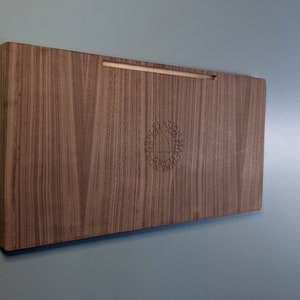 K&ouml;nnte beinhalten: Ein Wandregal aus Holz mit einer hellbraunen Holzmaserung. Das Regal hat ein kleines, eingraviertes Logo in der Mitte.