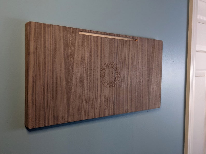 K&ouml;nnte beinhalten: Ein Wandregal aus Holz mit einer hellbraunen Holzmaserung. Das Regal hat ein kleines, eingraviertes Logo in der Mitte.
