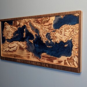 K&ouml;nnte beinhalten: Ein Wandkunstst&uuml;ck aus Holz, das das Mittelmeer darstellt. Das Meer ist mit blauem Harz gef&uuml;llt und das umliegende Land ist aus Holz. Das St&uuml;ck ist in einem dunklen Holzrahmen gerahmt und tr&auml;gt die Aufschrift "MEDITERRANEAN SEA" am unteren Rand.