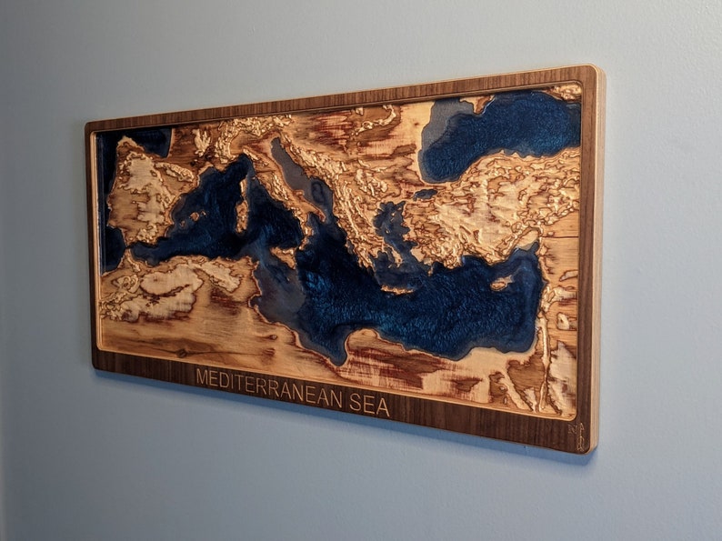 K&ouml;nnte beinhalten: Ein Wandkunstst&uuml;ck aus Holz, das das Mittelmeer darstellt. Das Meer ist mit blauem Harz gef&uuml;llt und das umliegende Land ist aus Holz. Das St&uuml;ck ist in einem dunklen Holzrahmen gerahmt und tr&auml;gt die Aufschrift "MEDITERRANEAN SEA" am unteren Rand.