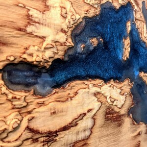 K&ouml;nnte beinhalten: Nahaufnahme einer Holzplatte mit einer blauen Harzeinlage. Das Harz ist in Form eines Flusses oder Sees geformt, und das Holz hat ein nat&uuml;rliches, rustikales Aussehen.