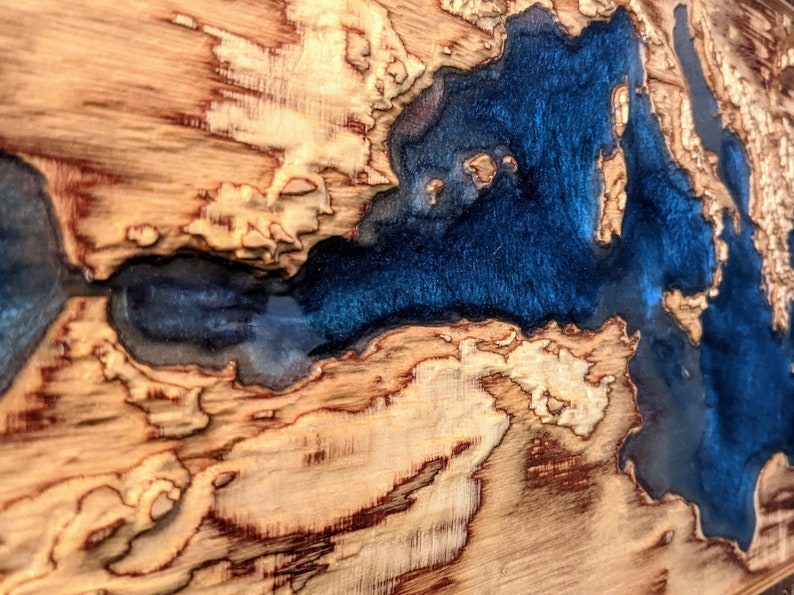 K&ouml;nnte beinhalten: Nahaufnahme einer Holzplatte mit einer blauen Harzeinlage. Das Harz ist in Form eines Flusses oder Sees geformt, und das Holz hat ein nat&uuml;rliches, rustikales Aussehen.