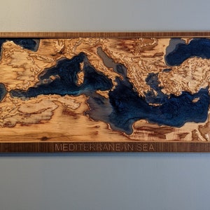 K&ouml;nnte beinhalten: Ein Wandkunstst&uuml;ck aus Holz, das das Mittelmeer darstellt. Das Meer ist mit blauem Harz gef&uuml;llt und das umgebende Land ist aus Holz. Das St&uuml;ck ist in einem dunklen Holzrahmen gerahmt und tr&auml;gt am unteren Rand den Text "MEDITERRANEAN SEA".