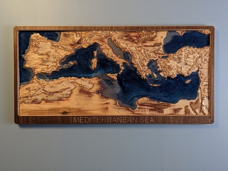 K&ouml;nnte beinhalten: Ein Wandkunstst&uuml;ck aus Holz, das das Mittelmeer darstellt. Das Meer ist mit blauem Harz gef&uuml;llt und das umgebende Land ist aus Holz. Das St&uuml;ck ist in einem dunklen Holzrahmen gerahmt und tr&auml;gt am unteren Rand den Text "MEDITERRANEAN SEA".