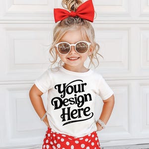 May include: A young girl wearing a white t-shirt with the text "Your Design Here" in black. She is wearing a red bow in her hair and white sunglasses. She is also wearing red and white polka dot shorts.