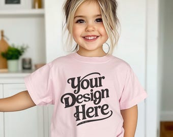 Puede incluir: Una camiseta rosa claro con el texto "Your Design Here" en negro, con una fuente retro. La camiseta de manga corta la lleva un niño pequeño con pelo rubio y un lazo rosa.