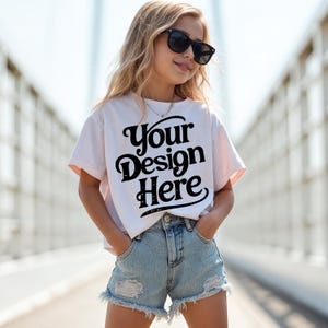 May include: A white t-shirt with the text "Your Design Here" in a black, stylized font. The shirt is worn with light blue denim shorts. The model is wearing black sunglasses.