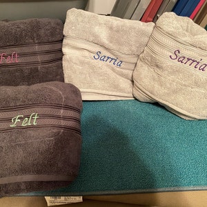 Op de afbeelding: Vier grijze handdoeken met geborduurde namen, "Felt" en "Sarria", op een turquoise oppervlak.
