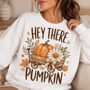 May include: A white long-sleeve sweatshirt featuring a graphic print of a wooden wagon carrying an orange pumpkin and a white daisy. The design includes autumn leaves, small hearts, and the brown text "HEY THERE PUMPKIN" printed above and below.