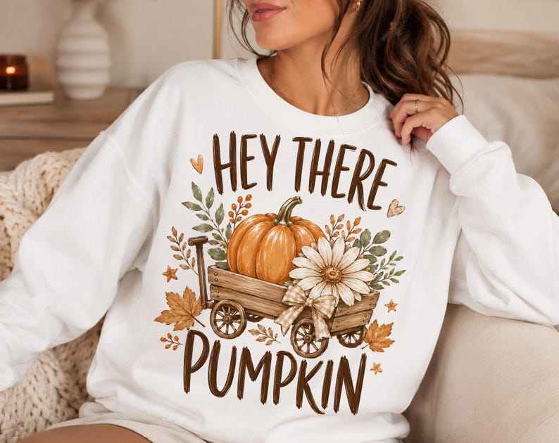 May include: A white long-sleeve sweatshirt featuring a graphic print of a wooden wagon carrying an orange pumpkin and a white daisy. The design includes autumn leaves, small hearts, and the brown text "HEY THERE PUMPKIN" printed above and below.