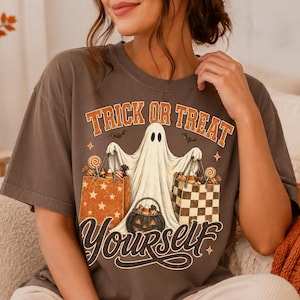 Op de afbeelding: Een donkergrijs t-shirt met korte mouwen en een afbeelding van een witte geest die twee boodschappentassen vol snoep en een pompoenemmer vasthoudt. De tekst luidt TRICK OR TREAT YOURSELF in oranje en witte gestileerde letters.