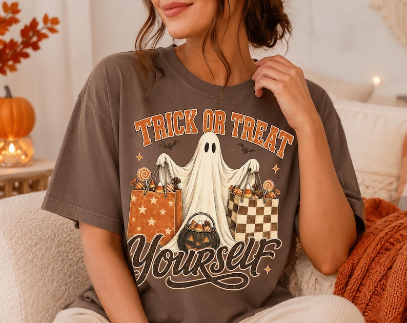 Op de afbeelding: Een donkergrijs t-shirt met korte mouwen en een afbeelding van een witte geest die twee boodschappentassen vol snoep en een pompoenemmer vasthoudt. De tekst luidt TRICK OR TREAT YOURSELF in oranje en witte gestileerde letters.