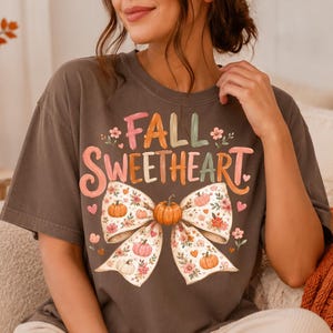May include: A dark gray short-sleeve t-shirt featuring the text FALL SWEETHEART in colorful letters above a large graphic of a bow. The bow is decorated with small pumpkins, flowers, and hearts in shades of orange, pink, and green.