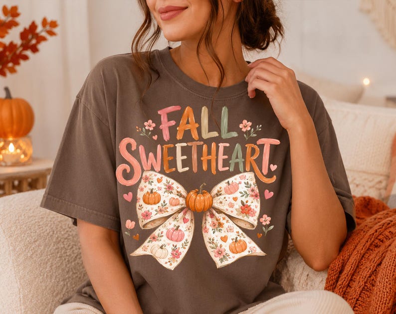 May include: A dark gray short-sleeve t-shirt featuring the text FALL SWEETHEART in colorful letters above a large graphic of a bow. The bow is decorated with small pumpkins, flowers, and hearts in shades of orange, pink, and green.