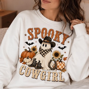 Op de afbeelding: Een wit sweatshirt met ronde hals en een grafische print van een geest met een zwarte cowboyhoed en een zwart-wit geblokte bandana. Het ontwerp bevat pompoenen, zonnebloemen, vleermuizen en een hoefijzer met de tekst SPOOKY COWGIRL.