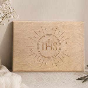 May include: A light-coloured wooden plaque featuring the engraved letters IHS within a sunburst design. The rectangular plaque has a natural wood grain. The design includes a cross above the letters and star accents.