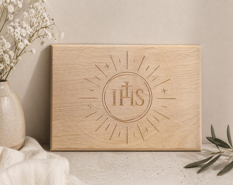 May include: A light-coloured wooden plaque featuring the engraved letters IHS within a sunburst design. The rectangular plaque has a natural wood grain. The design includes a cross above the letters and star accents.