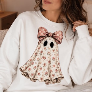 Puede incluir: Una sudadera blanca de manga larga con un gr&aacute;fico central de una figura de fantasma. El fantasma tiene un estampado floral peque&ntilde;o en tonos rosa y verde y lleva un lazo floral grande a juego en la parte superior. Tiene dos ojos ovalados negros.