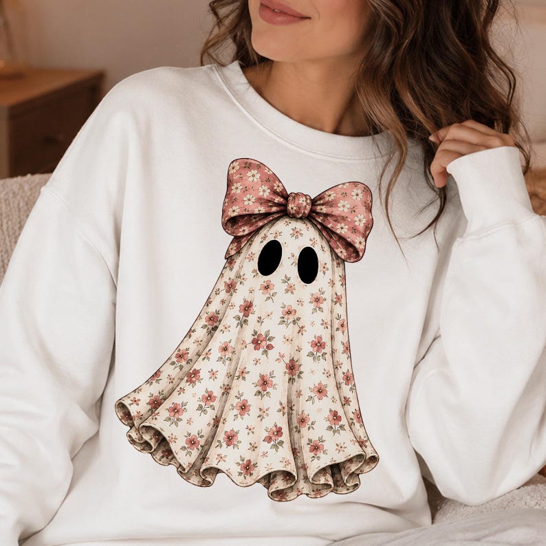 Puede incluir: Una sudadera blanca de manga larga con un gr&aacute;fico central de una figura de fantasma. El fantasma tiene un estampado floral peque&ntilde;o en tonos rosa y verde y lleva un lazo floral grande a juego en la parte superior. Tiene dos ojos ovalados negros.