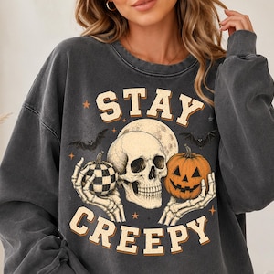 Op de afbeelding: Een antracietgrijs sweatshirt met ronde hals en een grafische print van een menselijke schedel die een pompoen met ruitjespatroon en een lantaarnpompoen vasthoudt. Het ontwerp bevat vleermuizen, een volle maan, kleine sterren en de tekst STAY CREEPY.