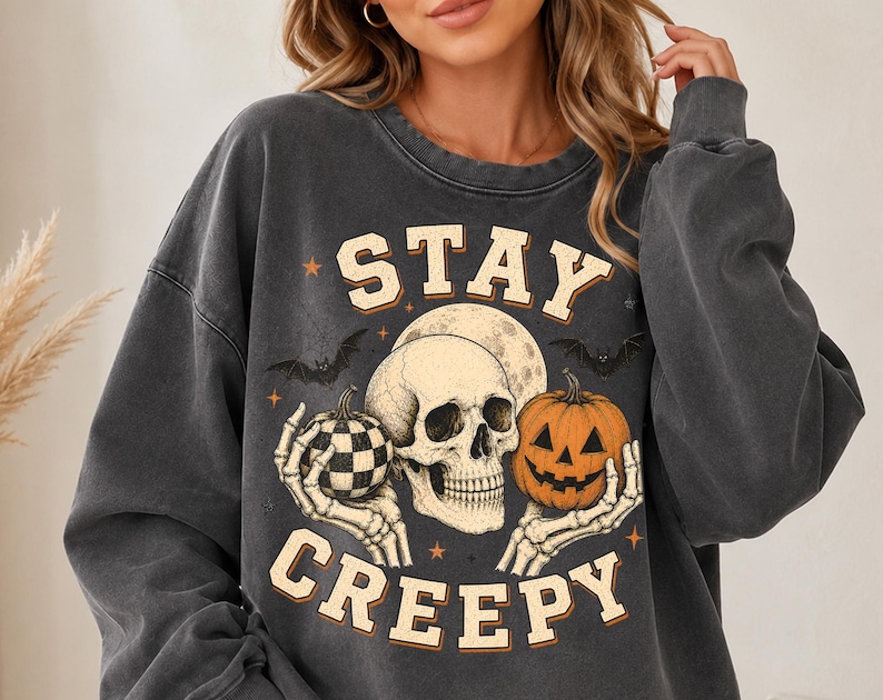 Op de afbeelding: Een antracietgrijs sweatshirt met ronde hals en een grafische print van een menselijke schedel die een pompoen met ruitjespatroon en een lantaarnpompoen vasthoudt. Het ontwerp bevat vleermuizen, een volle maan, kleine sterren en de tekst STAY CREEPY.
