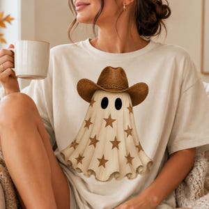 Puede incluir: Una camiseta de manga corta color crema con un gr&aacute;fico de un fantasma que lleva un sombrero de vaquero marr&oacute;n. El fantasma est&aacute; decorado con un patr&oacute;n de estrellas marrones y tiene dos ojos ovalados de color negro s&oacute;lido.