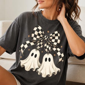 Op de afbeelding: Een antracietgrijs t-shirt met korte mouwen en een grafische print van twee witte geesten die aan een grote zwart-wit geblokte strik hangen. Het ontwerp bevat kleine beige steraccenten verspreid rond de geesten en de strik.