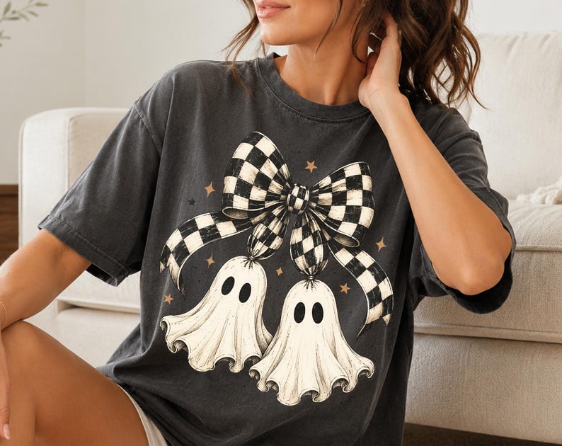 Op de afbeelding: Een antracietgrijs t-shirt met korte mouwen en een grafische print van twee witte geesten die aan een grote zwart-wit geblokte strik hangen. Het ontwerp bevat kleine beige steraccenten verspreid rond de geesten en de strik.