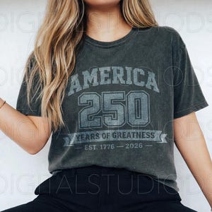 May include: Dark gray t-shirt with the text "AMERICA 250 YEARS OF GREATNESS EST. 1776 - 2026" in a distressed, vintage style. The shirt has a crew neck and short sleeves. The shirt is made of a soft, comfortable material.