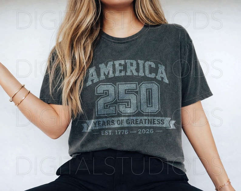 May include: Dark gray t-shirt with the text "AMERICA 250 YEARS OF GREATNESS EST. 1776 - 2026" in a distressed, vintage style. The shirt has a crew neck and short sleeves. The shirt is made of a soft, comfortable material.