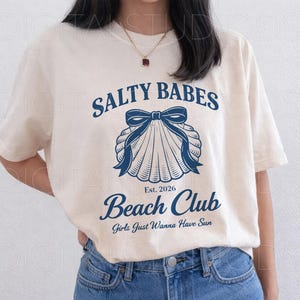 May include: Cream-colored t-shirt with the text "SALTY BABES Beach Club" in navy blue, featuring a seashell graphic with a bow. The shirt also includes the text "Est. 2026" and "Girls Just Wanna Have Sun".