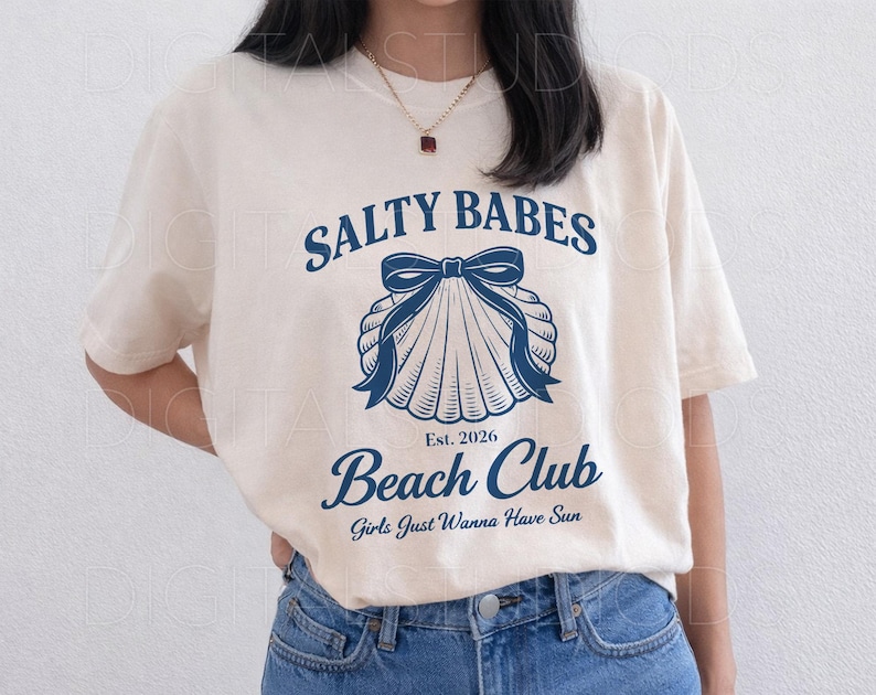 May include: Cream-colored t-shirt with the text "SALTY BABES Beach Club" in navy blue, featuring a seashell graphic with a bow. The shirt also includes the text "Est. 2026" and "Girls Just Wanna Have Sun".