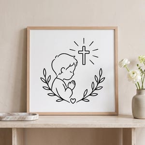 May include: A framed black and white print depicting a child in prayer, with a cross above. The child is encircled by a wreath of leaves and a heart. The artwork is displayed on a light-coloured wooden shelf.