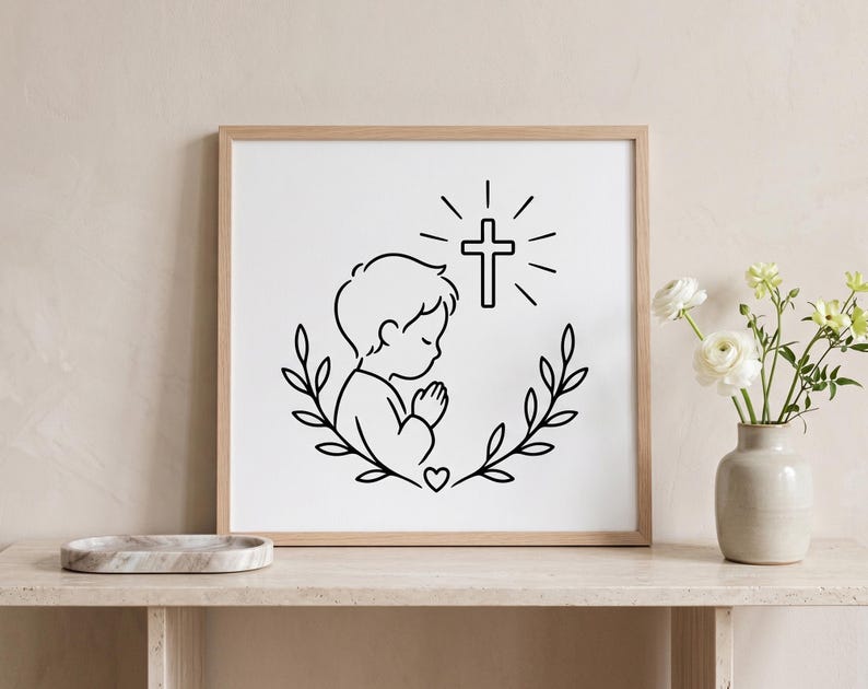 May include: A framed black and white print depicting a child in prayer, with a cross above. The child is encircled by a wreath of leaves and a heart. The artwork is displayed on a light-coloured wooden shelf.