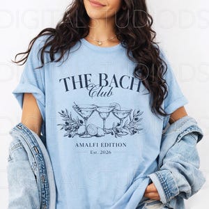 May include: Light blue t-shirt with the text "THE BACH CLUB" above a graphic of three martini glasses, olives, and lemons. The text "AMALFI EDITION Est. 2026" is below the graphic. The shirt is worn with a denim jacket.