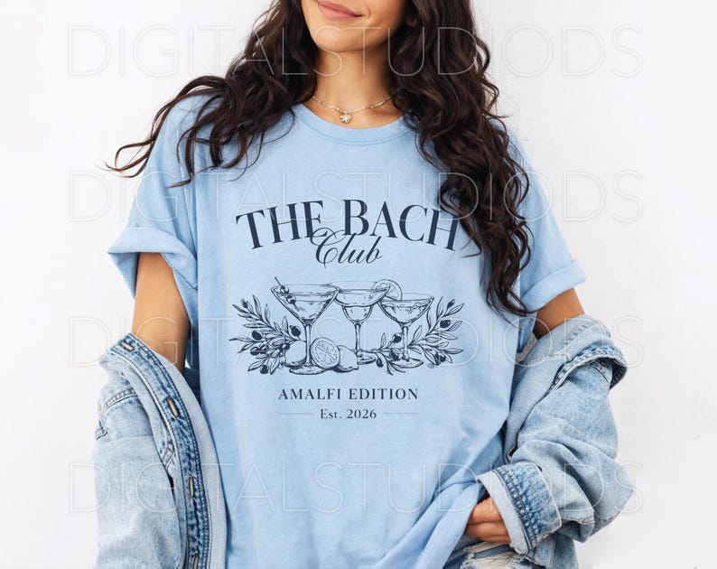 May include: Light blue t-shirt with the text "THE BACH CLUB" above a graphic of three martini glasses, olives, and lemons. The text "AMALFI EDITION Est. 2026" is below the graphic. The shirt is worn with a denim jacket.