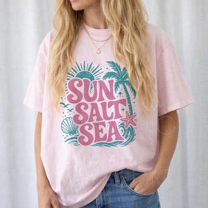 May include: A light pink t-shirt with the words "SUN SALT SEA" in a retro font. The design includes a sun, palm tree, seashell, starfish, and waves in teal and pink. The shirt is a relaxed fit.