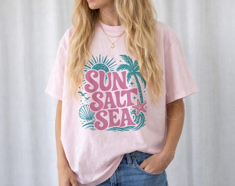 May include: A light pink t-shirt with the words "SUN SALT SEA" in a retro font. The design includes a sun, palm tree, seashell, starfish, and waves in teal and pink. The shirt is a relaxed fit.