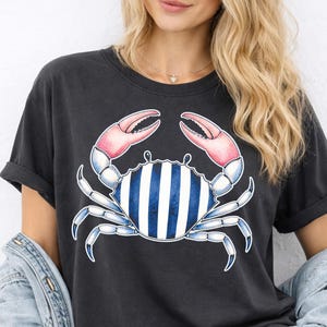 May include: Dark gray t-shirt featuring a blue crab illustration with pink claws and a striped body. The crab design is centered on the shirt. The shirt has short sleeves and a relaxed fit.