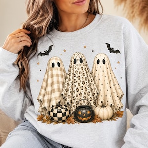 Op de afbeelding: Een lichtgrijs sweatshirt met ronde hals en een afbeelding van drie geesten achter pompoenen. De geesten hebben patronen van beige ruitjes, luipaardprint en zwarte sterren. Twee zwarte vleermuizen en kleine gouden sterren zijn boven de geesten gedrukt.