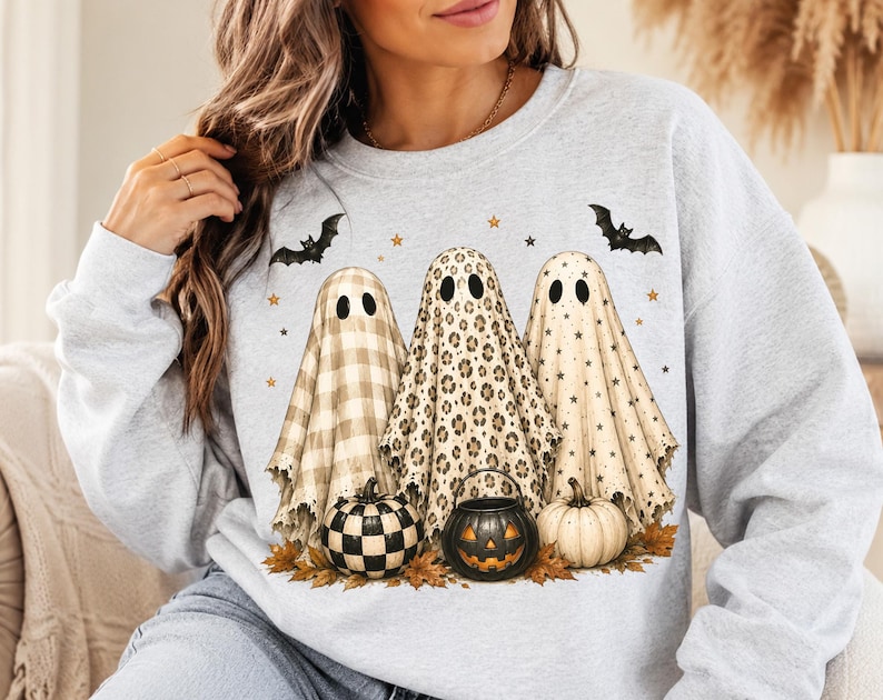 Op de afbeelding: Een lichtgrijs sweatshirt met ronde hals en een afbeelding van drie geesten achter pompoenen. De geesten hebben patronen van beige ruitjes, luipaardprint en zwarte sterren. Twee zwarte vleermuizen en kleine gouden sterren zijn boven de geesten gedrukt.