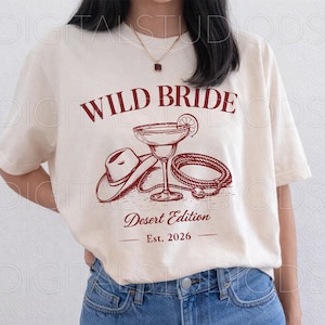 May include: Cream-colored t-shirt with a maroon graphic that reads "WILD BRIDE Desert Edition Est. 2026." The graphic features a cowboy hat, margarita glass, and rope. The shirt is a relaxed fit.