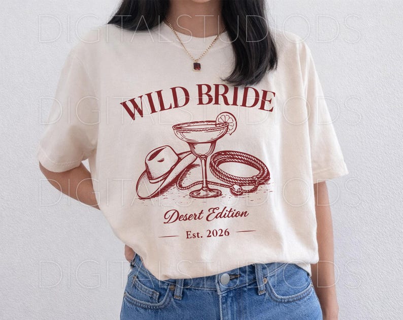 May include: Cream-colored t-shirt with a maroon graphic that reads "WILD BRIDE Desert Edition Est. 2026." The graphic features a cowboy hat, margarita glass, and rope. The shirt is a relaxed fit.
