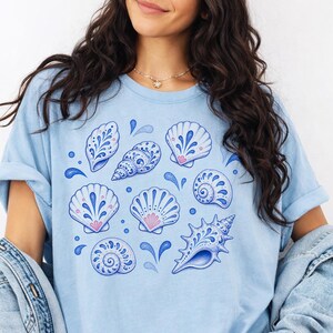 May include: Light blue t-shirt featuring a collection of stylized seashells in shades of blue and pink. The design includes various shell shapes with intricate patterns and water droplet accents, creating a coastal theme.