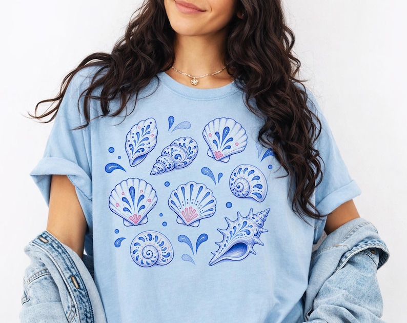 May include: Light blue t-shirt featuring a collection of stylized seashells in shades of blue and pink. The design includes various shell shapes with intricate patterns and water droplet accents, creating a coastal theme.